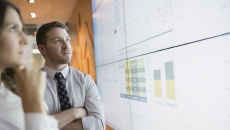 Two people standing up and looking at a large screen with graphs on it Two people standing up and looking at a large screen with graphs on it