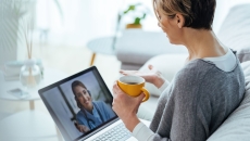 Person sitting on a couch speaking to a healthcare professional on a computer while holding a cup in their hand Person sitting on a couch speaking to a healthcare professional on a computer while holding a cup in their hand