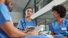 Four healthcare professionals wearing blue scrubs and sitting in a circle with one holding a tablet Four healthcare professionals wearing blue scrubs and sitting in a circle with one holding a tablet