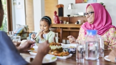 Three people sitting around a table with food on it. One person is wearing a pink head scarf, one a blue shirt and the other a white shirt Three people sitting around a table with food on it. One person is wearing a pink head scarf, one a blue shirt and the other a white shirt