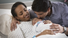 A person laying in a hospital bed with a newborn on their lap and another person standing beside them A person laying in a hospital bed with a newborn on their lap and another person standing beside them