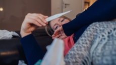 Person lying on their couch looking at a thermometer Person lying on their couch looking at a thermometer