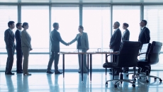 Group of three people on left, two people shaking hands in middle, three people standing behind office chairs on right Group of three people on left, two people shaking hands in middle, three people standing behind office chairs on right
