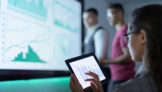 Person sitting while looking at a tablet with two other people standing up looking at a graph on a screen Person sitting while looking at a tablet with two other people standing up looking at a graph on a screen