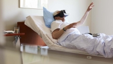 Person in a clinical setting lying in bed with their hand raised while wearing a virtual reality headset