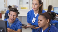 Three healthcare providers wearing scrubs sitting around a computer looking at the screen Three healthcare providers wearing scrubs sitting around a computer looking at the screen