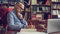 Person sitting at a desk writing something down on a piece of paper with a bookshelf behind them Person sitting at a desk writing something down on a piece of paper with a bookshelf behind them