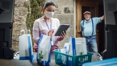 A woman using a tablet delivering medications while a man stands in his doorway in the background A woman using a tablet delivering medications while a man stands in his doorway in the background