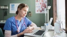 Healthcare provider wearing a stethoscope around their neck and scrubs sitting at a desk and looking at a computer