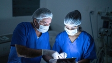 Two healthcare providers in scrubs wearing masks while in a dark room looking at a tablet