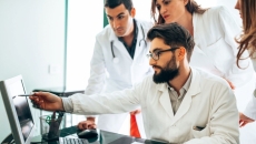 Three people wearing lab coats with one seated and pointing at screen