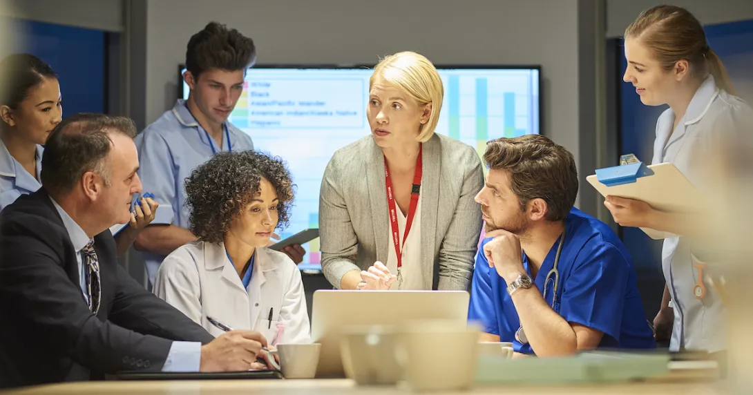 Several people standing and sitting around a table with a screen in the background that has a graph on it Several people standing and sitting around a table with a screen in the background that has a graph on it