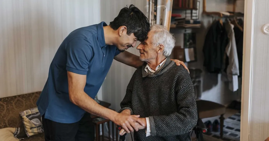 Two individuals with one in a wheelchair and the other standing over them with the two putting their heads together Two individuals with one in a wheelchair and the other standing over them with the two putting their heads together