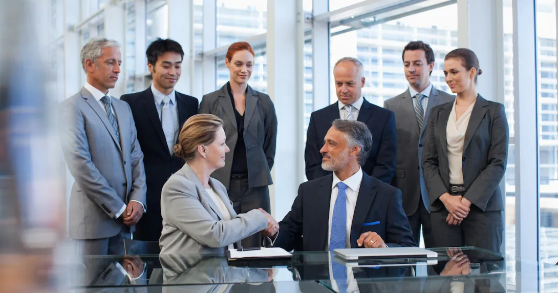 Several business people standing around a table watching two people sitting down shaking hands Several business people standing around a table watching two people sitting down shaking hands