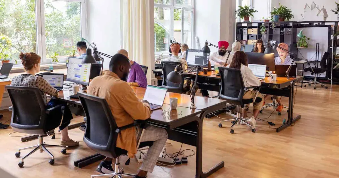 Numerous employees sitting at desks in a room with large windows Numerous employees sitting at desks in a room with large windows