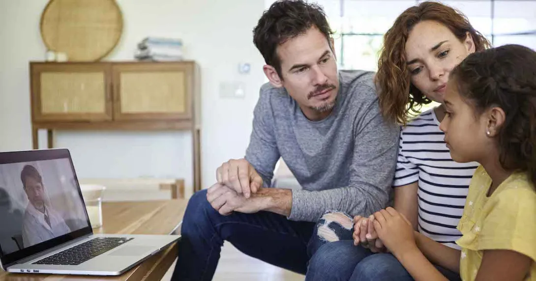 Three people sitting on a couch with a computer on a coffee table showing a healthcare provider on the screen Three people sitting on a couch with a computer on a coffee table showing a healthcare provider on the screen