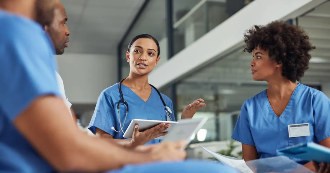 Four healthcare professionals wearing blue scrubs and sitting in a circle with one holding a tablet Four healthcare professionals wearing blue scrubs and sitting in a circle with one holding a tablet