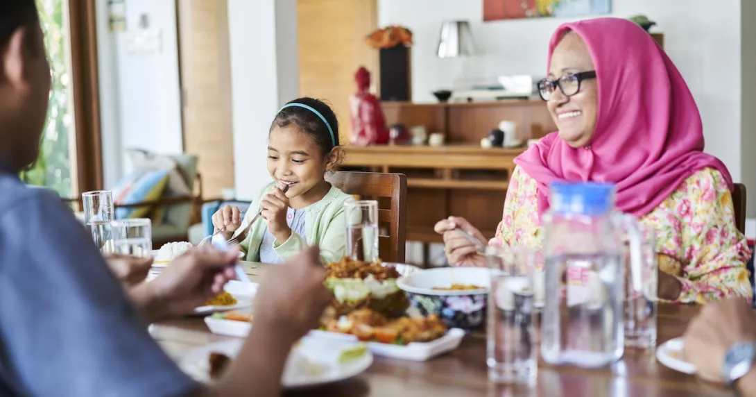 Three people sitting around a table with food on it. One person is wearing a pink head scarf, one a blue shirt and the other a white shirt Three people sitting around a table with food on it. One person is wearing a pink head scarf, one a blue shirt and the other a white shirt