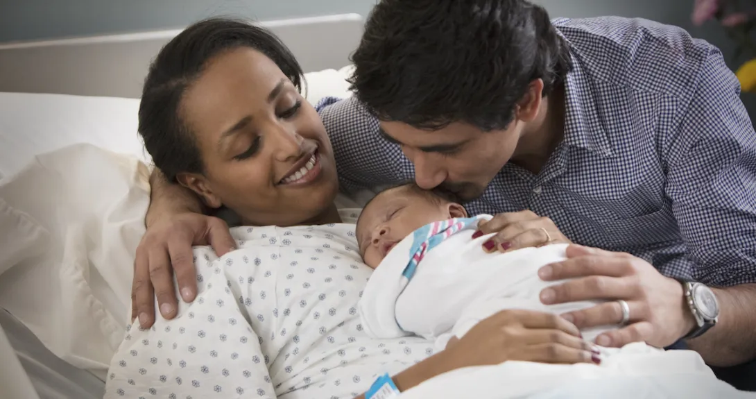 A person laying in a hospital bed with a newborn on their lap and another person standing beside them A person laying in a hospital bed with a newborn on their lap and another person standing beside them