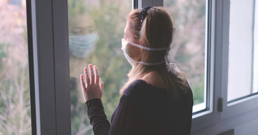 Person with a mask on standing by a window looking out Person with a mask on standing by a window looking out