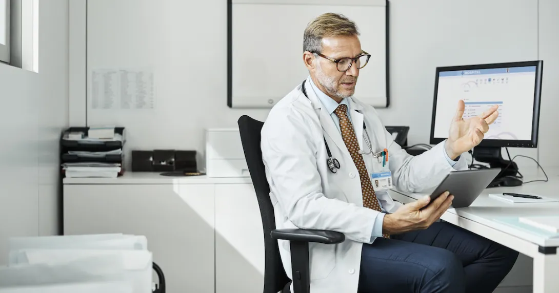 Healthcare provider sitting in a chair at a desk with a computer on it while looking at a tablet Healthcare provider sitting in a chair at a desk with a computer on it while looking at a tablet
