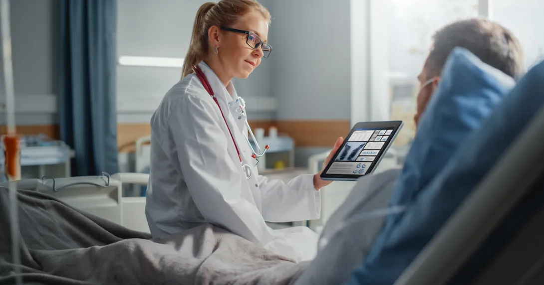 Healthcare provider sitting on a patient's hospital bed while holding a tablet showing diagnostic images of lungs Healthcare provider sitting on a patient's hospital bed while holding a tablet showing diagnostic images of lungs