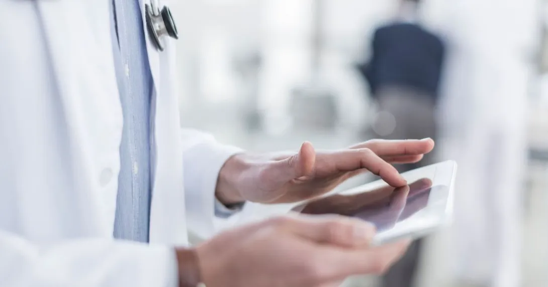 The body of a healthcare provider using a tablet while wearing a lab coat and stethoscope around their neck The body of a healthcare provider using a tablet while wearing a lab coat and stethoscope around their neck