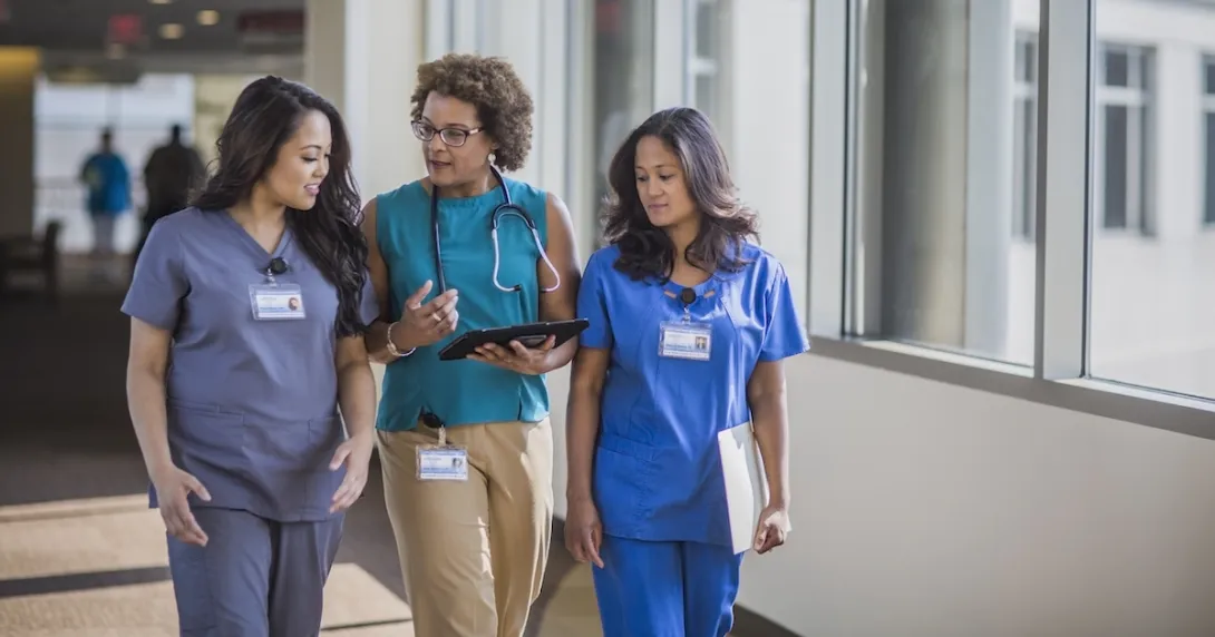 Three people walking down a hall, two healthcare providers in scrubs and one person in civilian clothing Three people walking down a hall, two healthcare providers in scrubs and one person in civilian clothing