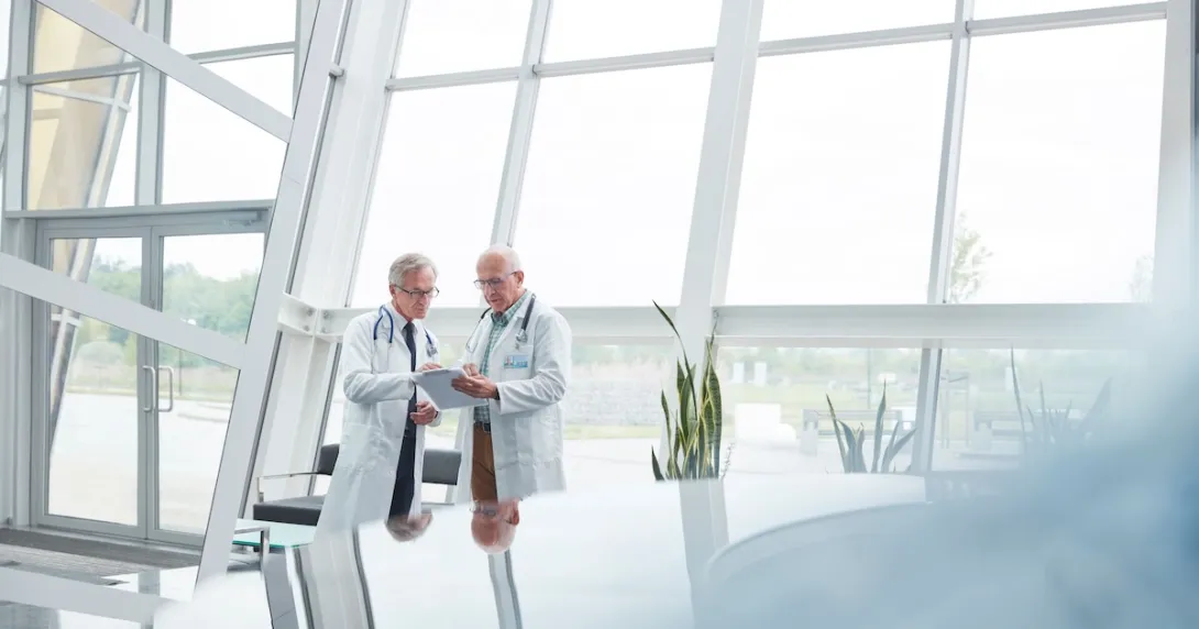 Two healthcare providers standing in a room in front of large windows Two healthcare providers standing in a room in front of large windows