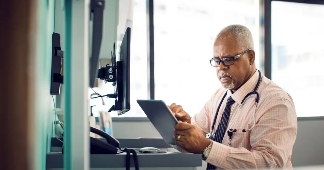 "Healthcare professional sitting at a desk with a window behind them while wearing a stethoscope around their neck and looking at a tablet" "Healthcare professional sitting at a desk with a window behind them while wearing a stethoscope around their neck and looking at a tablet"