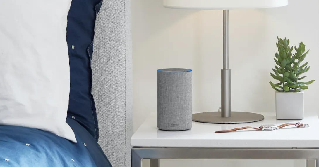 Light gray Amazon Alexa device on a bedside table next to a lamp, watch and houseplant