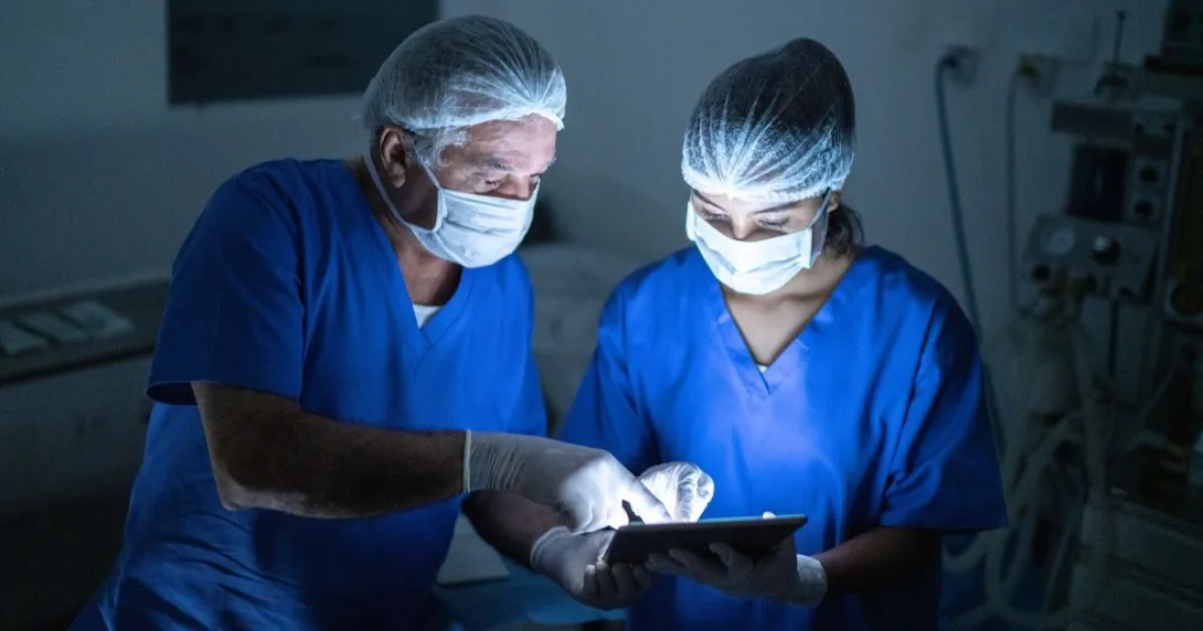 Two people wearing scrubs and masks in a procedure room look at a tablet. Two people wearing scrubs and masks in a procedure room look at a tablet.