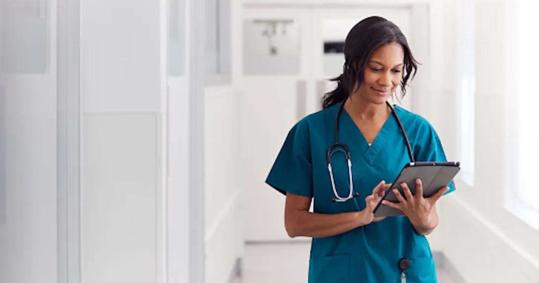 Nurse walking down a white hallway while wearing scrubs and looking at a tablet Nurse walking down a white hallway while wearing scrubs and looking at a tablet