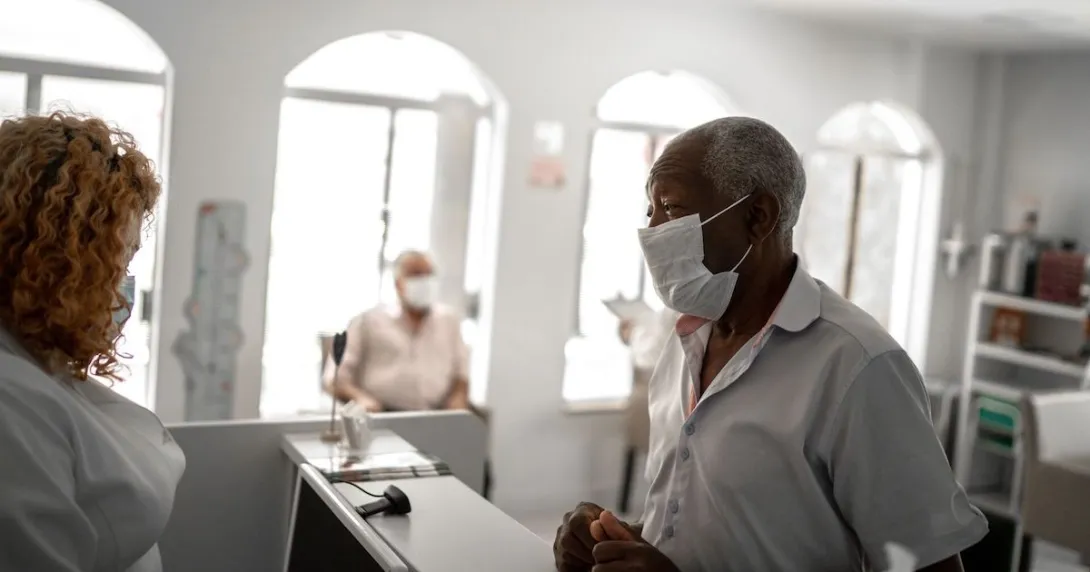 An older patient wearing a mask checking in a with a receptionist An older patient wearing a mask checking in a with a receptionist