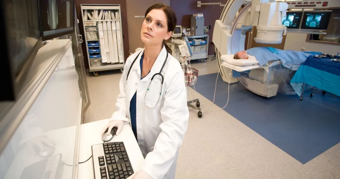 A medical professional looks at images while a patient receives a CT scan in the background A medical professional looks at images while a patient receives a CT scan in the background