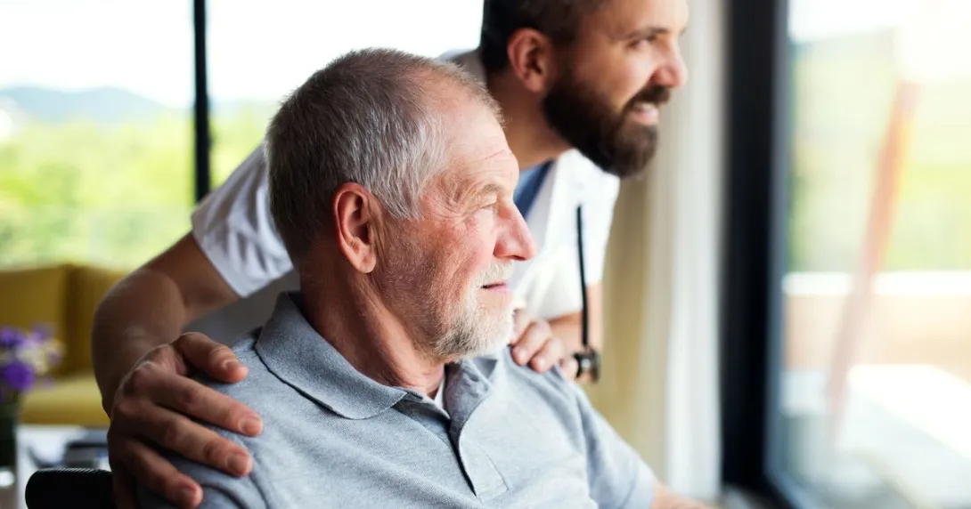 Person sitting in a wheelchair with a healthcare provider next to them, looking out a window Person sitting in a wheelchair with a healthcare provider next to them, looking out a window