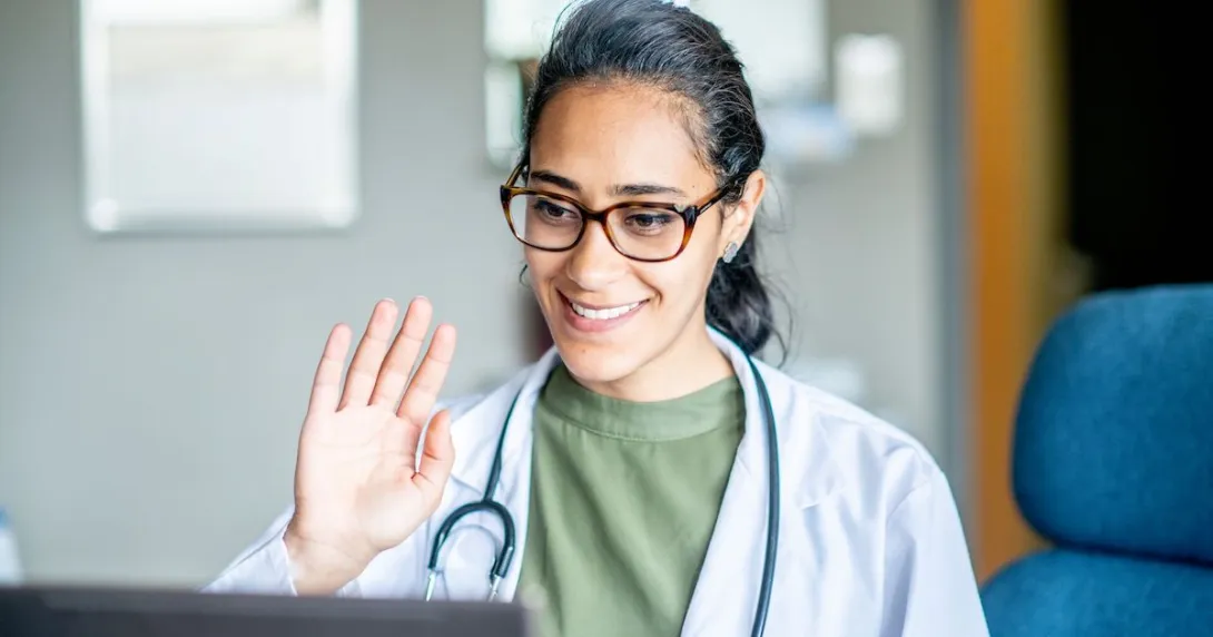 Healthcare provider sitting in front of a computer waving at the screen Healthcare provider sitting in front of a computer waving at the screen