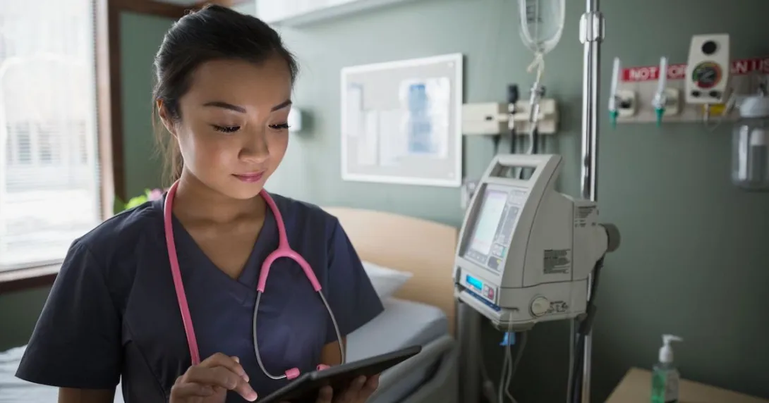 Healthcare provider standing in a hospital room wearing a pink stethoscope while holding a tablet Healthcare provider standing in a hospital room wearing a pink stethoscope while holding a tablet