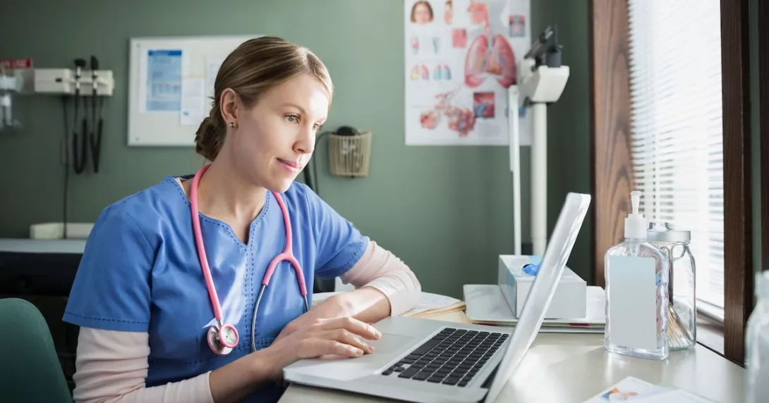 Healthcare provider in blue scrubs sitting at a desk looking at a computer Healthcare provider in blue scrubs sitting at a desk looking at a computer
