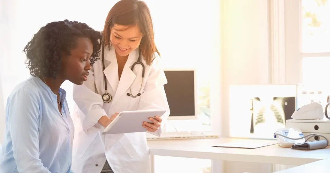 Healthcare provider standing over a patient sitting down while showing them a tablet Healthcare provider standing over a patient sitting down while showing them a tablet