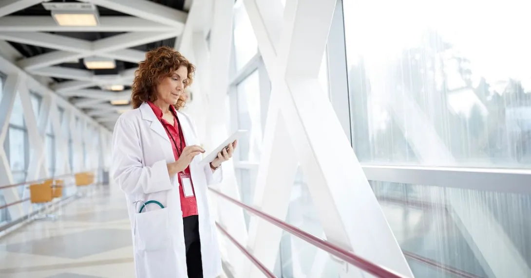 Healthcare provider standing in a hallway looking at a tablet next to a giant window