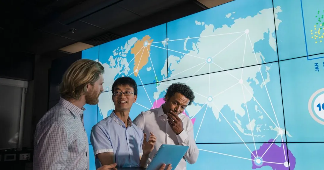 Three people standing in front of a screen showing continents connected by lines Three people standing in front of a screen showing continents connected by lines