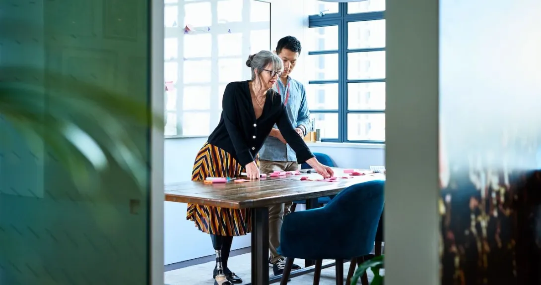 Two people standing in a room looking at something on a desk while standing above it