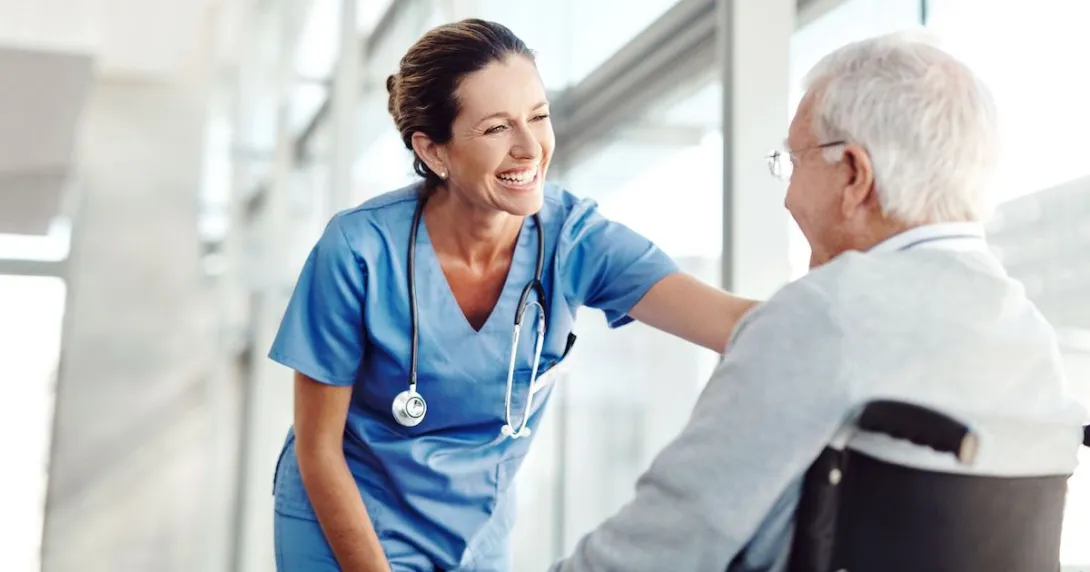 A nurse smiling and standing in front of a window leaning over a patient in a wheelchair A nurse smiling and standing in front of a window leaning over a patient in a wheelchair