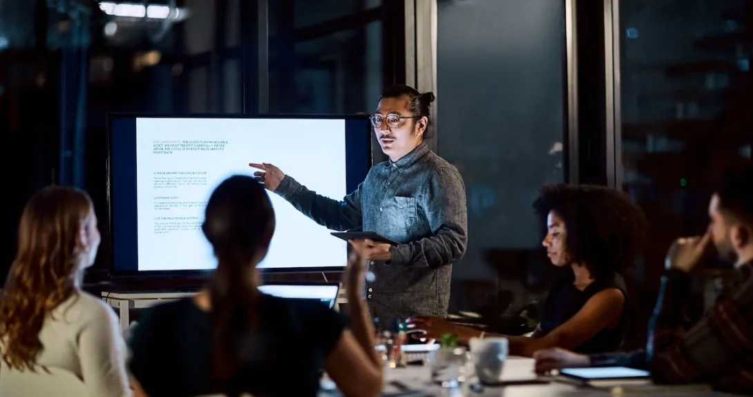Person presenting in front of other people while standing next to a screen in a dark room Person presenting in front of other people while standing next to a screen in a dark room