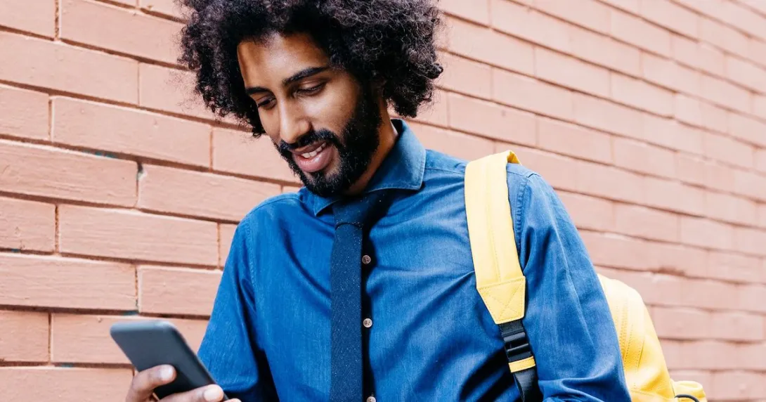 Person walking beside a brick wall wearing a blue shirt and yellow backpack while looking at a phone Person walking beside a brick wall wearing a blue shirt and yellow backpack while looking at a phone