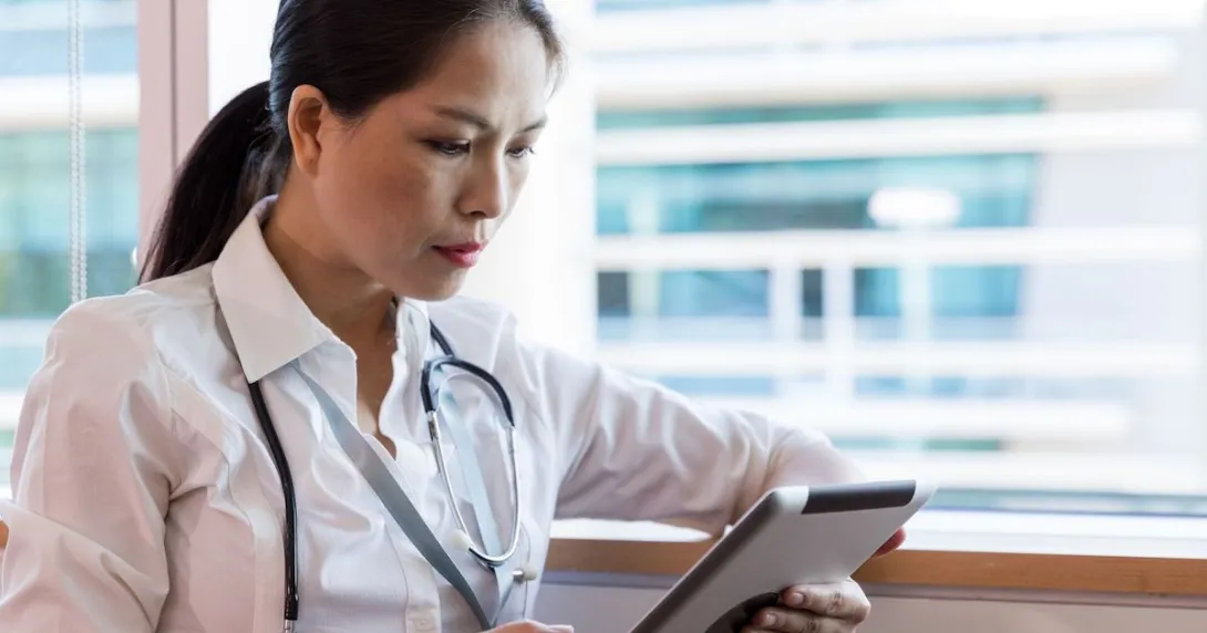 Healthcare provider sitting next to a window wearing a stethoscope around their neck and looking at a tablet Healthcare provider sitting next to a window wearing a stethoscope around their neck and looking at a tablet