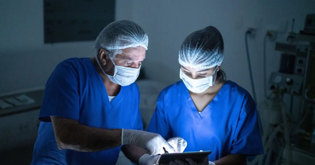 Two healthcare providers in scrubs wearing masks while in a dark room looking at a tablet Two healthcare providers in scrubs wearing masks while in a dark room looking at a tablet
