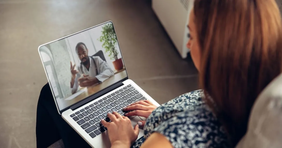 Person sitting on their couch with a computer on their lap talking to a virtual healthcare provider Person sitting on their couch with a computer on their lap talking to a virtual healthcare provider