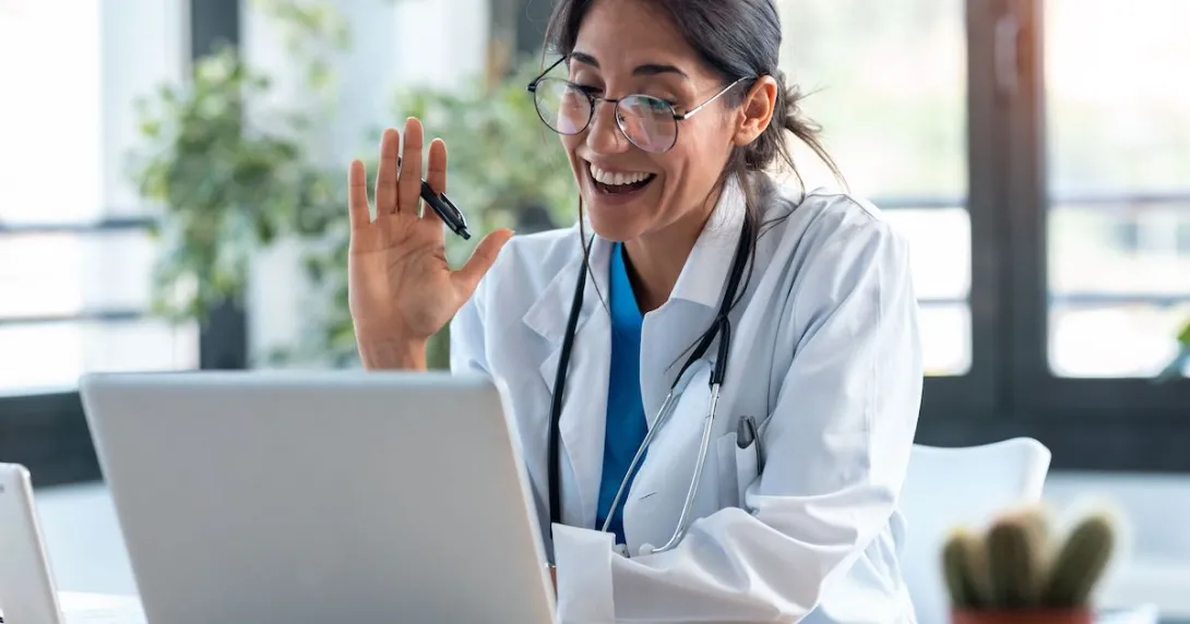 A healthcare provider in a lab coat with a stethoscope around their neck sitting at a desk looking at a computer and waving A healthcare provider in a lab coat with a stethoscope around their neck sitting at a desk looking at a computer and waving